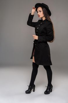 Full length portrait of a charming professional model posing sideways at studio against a gray background. She is weared in a checkered dress, black coat, hat, stockings and boots. Fashion shot. Sincere emotions concept.