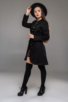 Full length portrait of a beautiful lady holding her hat and posing at studio against a gray background. She is weared in a checkered dress, black coat, stockings and boots. Fashion shot. Sincere emotions concept.