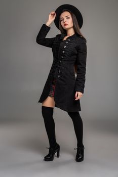 Full length portrait of a beautiful professional model posing at studio against a gray background. She is weared in a checkered dress, black coat, hat, stockings and boots. Fashion shot. Sincere emotions concept.