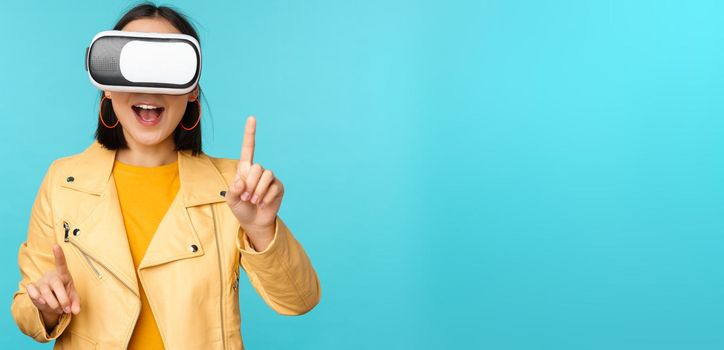 Young asian woman using virtual reality glasses, using VR headset, standing amused against blue background. Copy space