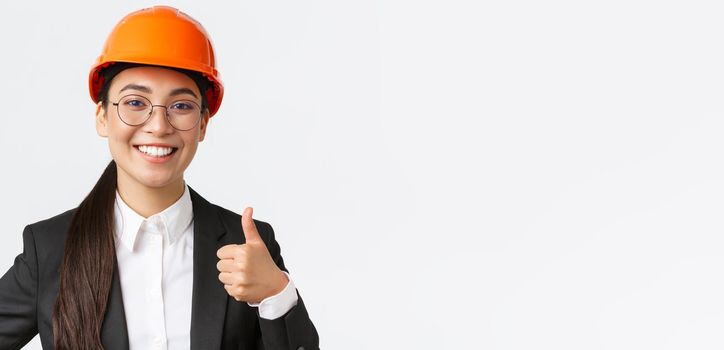Close-up of professional pleased asian female chief engineer, architect in business suit and safety helmet showing thumbs-up in approval, give permission, recommend construction company.