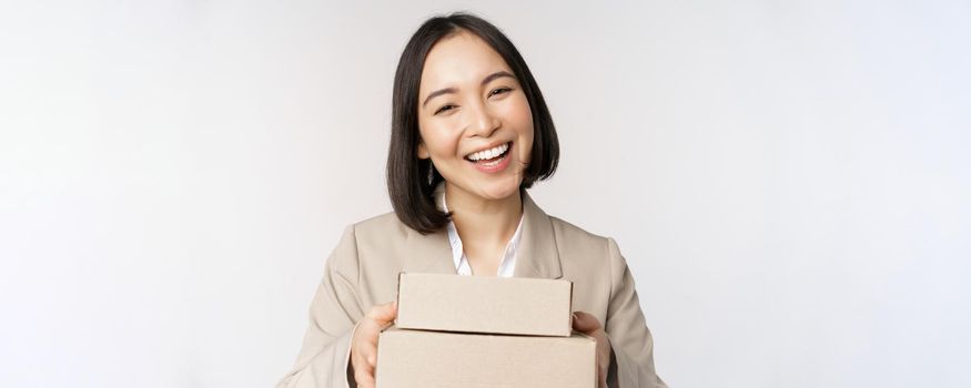 Image of asian saleswoman, business woman giving boxes with order, deliver to customer, standing in suit over white background.