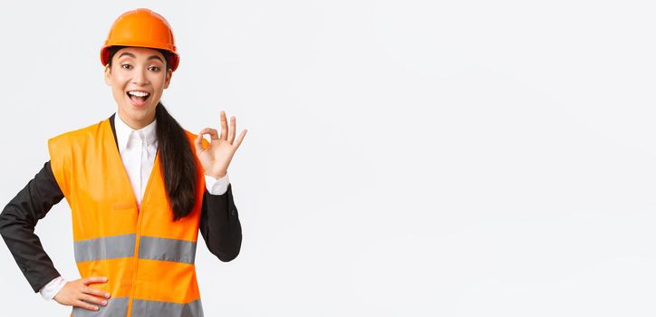 Satisfied happy asian female engineer, construction manager in safety helmet and reflective jacket showing okay gesture pleased, guarantee building quality, ensure everything excellent.