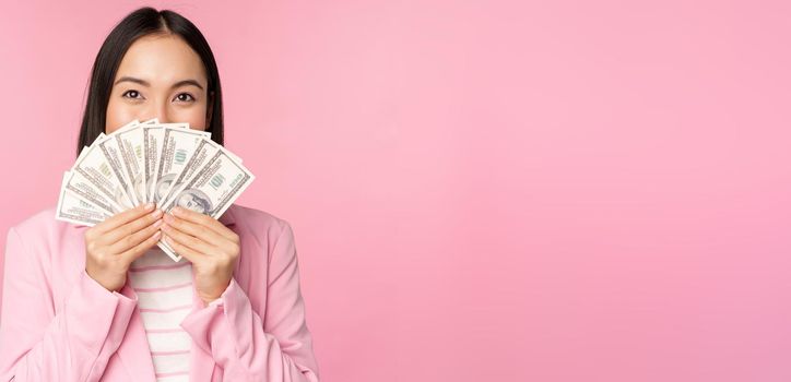 Happy asian lady in suit holding money, dollars with pleased face expression, standing over pink background. Copy space