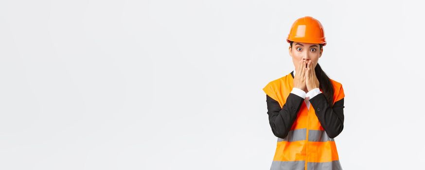 Concerned and worried asian female engineer having problem at construction area, gasping and cover mouth shocked, stare at camera startled with alarmed expression, wearing safety helmet.