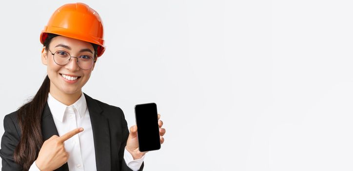 Close-up of smiling professional female asian construction engineer, architect in suit and safety mask introduce application, pointing finger at smartphone display, white background.