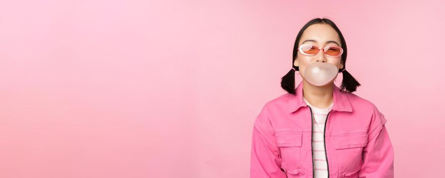 Stylish asian girl blowing bubblegum bubble, chewing gum, wearing sunglasses, posing against pink background. Copy space