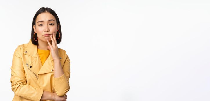 Image of stylish japanese girl looking with unamused, tired and upset face expression, standing in casual clothes over white background. Copy space