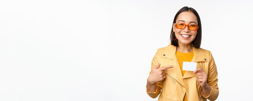 Portrait of beautiful modern asian girl in sunglasses, smiling happy, showing credit card, standing over white background. copy space