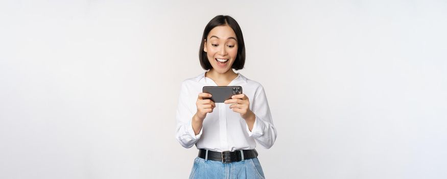 Image of young asian woman watching on smartphone app, holding mobile phone and looking at screen, laughing and smiling, standing over white background.