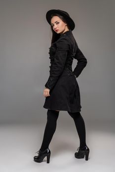 Full length portrait of an attractive girl looking at the camera and posing back at studio against a gray background. She is weared in a checkered dress, black coat, hat, stockings and boots. Fashion shot. Sincere emotions concept.