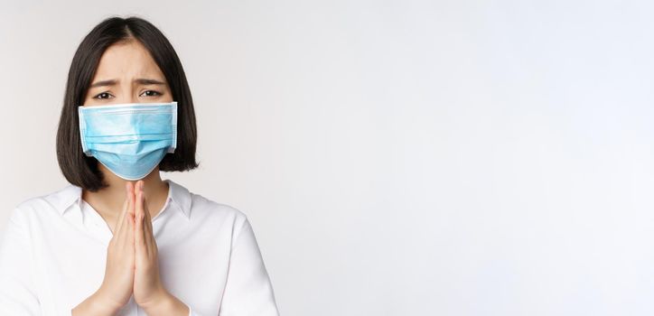 Portrait of asian woman in medical face mask from covid, begging, asking for help, say please, standing over white background.