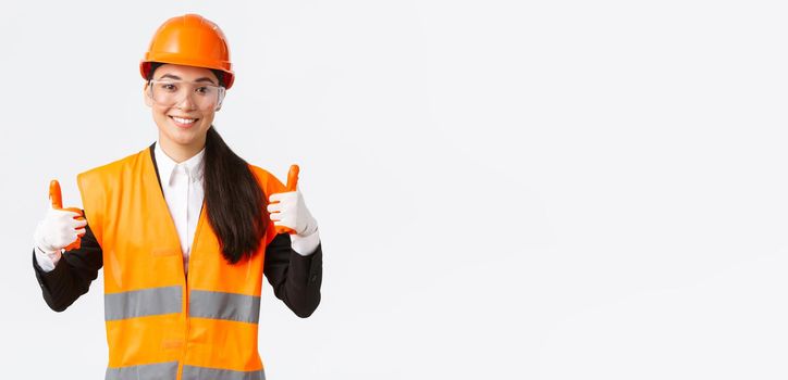 Satisfied smiling female asian chief construction engineer giving permission to enter building or entertprise after wearing safety clothing, glasses gloves and helmet, showing thumbs-up in approval.