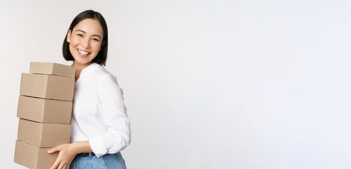 Portrait of young asian woman holding boxes, carry delivery goods. Korean female entrepreneur assemble order, standing voer white background.