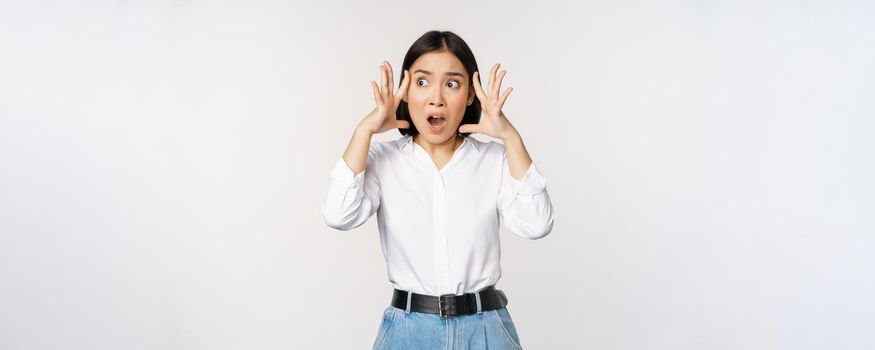 Image of shocked anxious asian woman in panic, holding hands on head and worrying, standing frustrated and scared against white background.