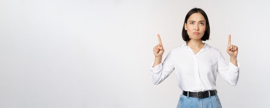 Skeptical businesswoman, asian office manager, pointing fingers up and grimacing doubtful, hesitating, standing over white background.