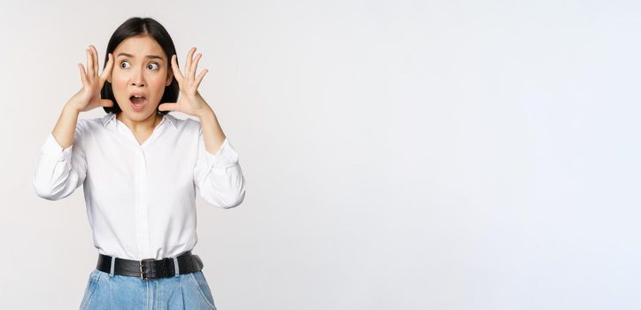 Image of shocked anxious asian woman in panic, holding hands on head and worrying, standing frustrated and scared against white background.