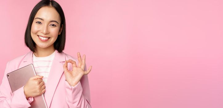 Portrait of corporate woman, girl in office in business suit, holding digital tablet, showing okay, recommending company, standing over pink background.