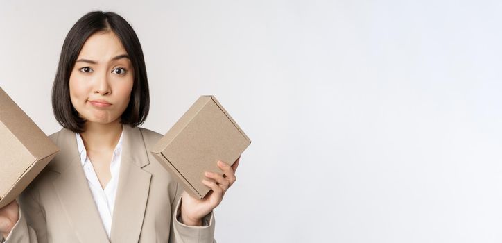 Puzzled asian saleswoman holding boxes, looking confused, standing in business suit over white background. Copy space