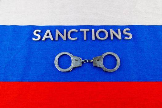 the word sanctions laid with silver metal letters on russian tricolor flag near handcuffs over it in directly above view