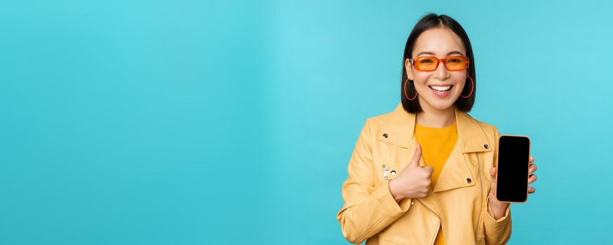 Young stylish chinese woman showing mobile phone screen and thumbs up, smartphone app, standing over blue background. copy space