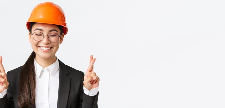 Close-up of hopeful optimistic asian female engineer, architect having faith in project, wearing safety helmet and business suit awaiting good results, cross fingers good luck, white background.