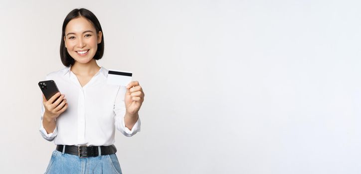 Online shopping concept. Smiling modern asian girl shows her credit card, holds mobile phone, order with smartphone, standing over white background.