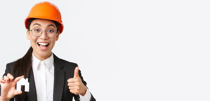 Close-up of upbeat professional female asian engineer, architect in helmet and business suit encourage contact her company for renovation and design, showing house and thumbs-up.