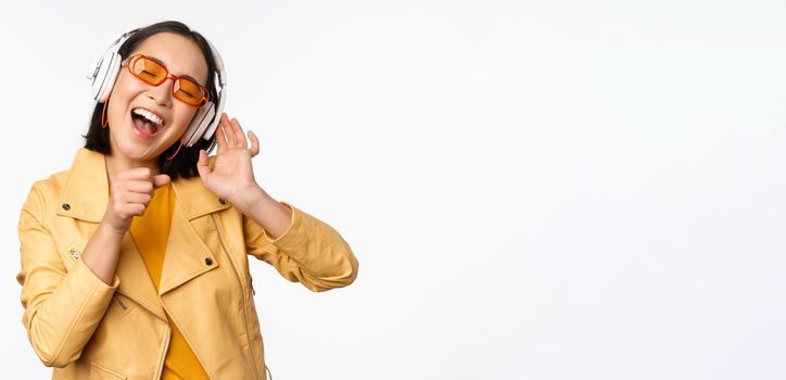 Beautiful asian girl, laughing happy, listening music in headphones, standing over white background. Copy space