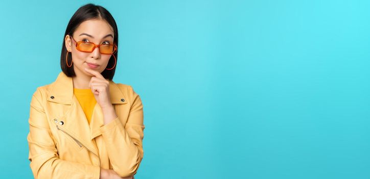 Portrait of asian woman thinking, looking thoughtful, searching ideas or solution, wearing sunglasses, standing over blue background.