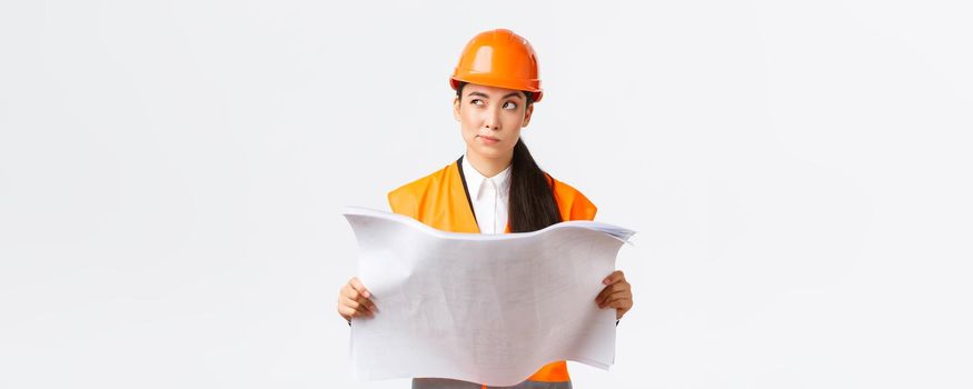 Thoughtful professional asian female construction manager, architect reading blueprints and looking away pondering, thinking, making decision while studying project plan, white background.