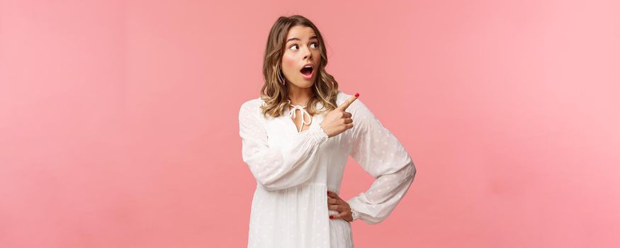 Surprised and excited good-looking blond girl in white cute dress, open mouth in amazement, look curious pointing upper right corner astonished, see something awesome, pink background.