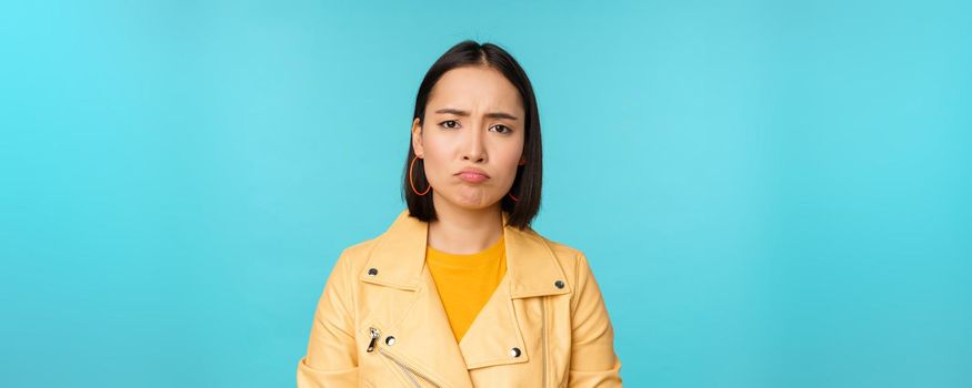 Close up of disappointed asian girl sulking, grimacing and frowning displeased, standing moody against blue background. Copy space