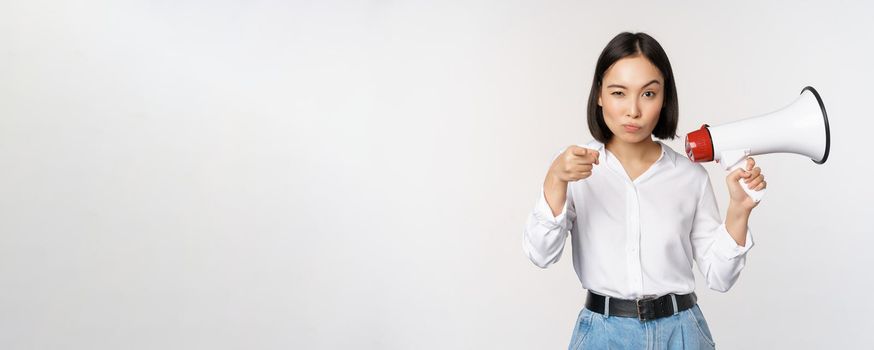 Image of modern asian woman with megaphone, pointing at you camera, making announcement, white background.