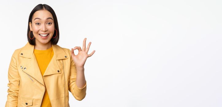 Happy beautiful asian girl shows okay, ok recommending sign, pleased by smth, praising quality, standing over white background. Copy space