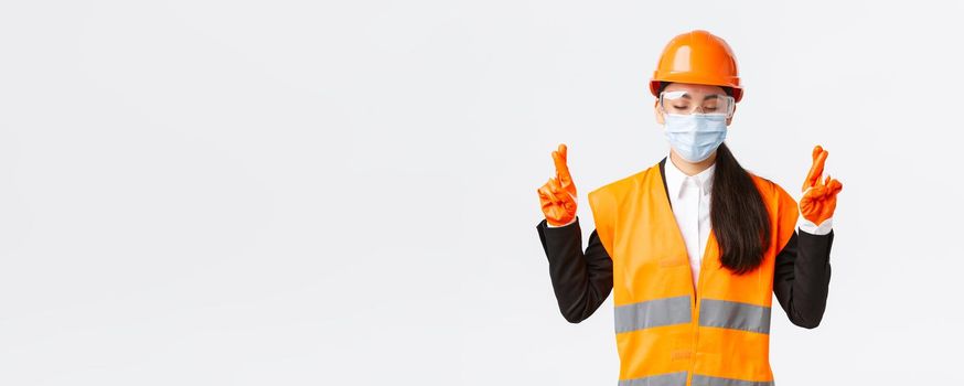Covid-19 safety protocol at enterpise, construction and preventing virus concept. Hopeful asian female engineer in helmet, face mask praying, close eyes and cross fingers good luck, white background.
