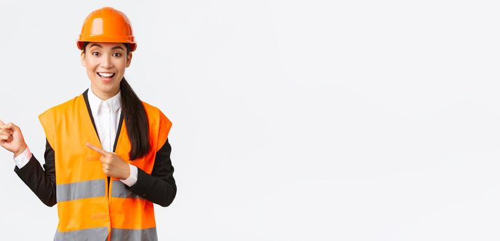 Building, construction and industrial concept. Smiling female engineer in helmet and reflective clothing showing way, pointing upper left corner, talking about constructing project, white background.