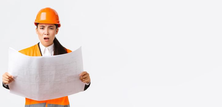 Frustrated and displeased female asian engineer, chief architect at construction zone, looking disappointed after studying blueprints, stare camera, scolding manager, white background.