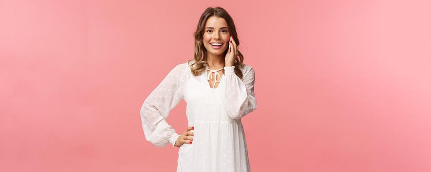 Portrait of joyful good-looking blond woman in white dress, talking on phone, hold smartphone near ear and look upbeat camera, smiling discuss friends date, stand pink background.