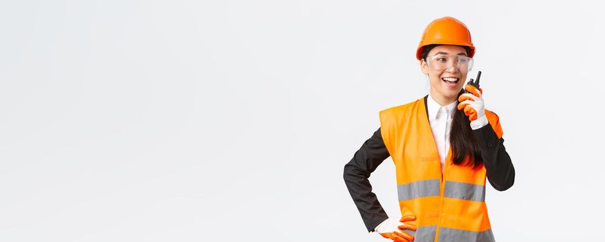 Confident pleased asian female engineer in safety helmet and uniform talking with chief architect using walkie-talkie. Satisfied construction technician contact team using radio phone.