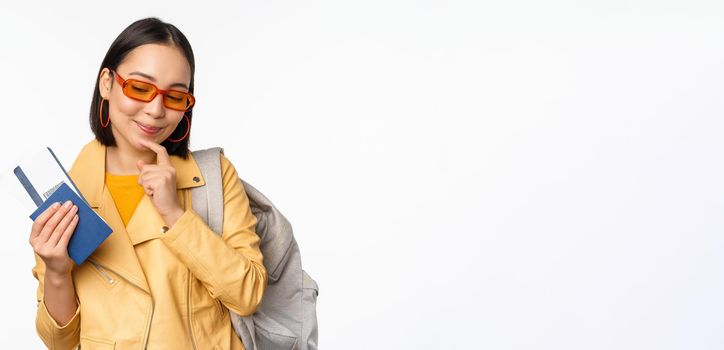 Asian girl tourist with boarding tickets and passport going abroad, holding backpack, thinking of travelling, standing over white background.