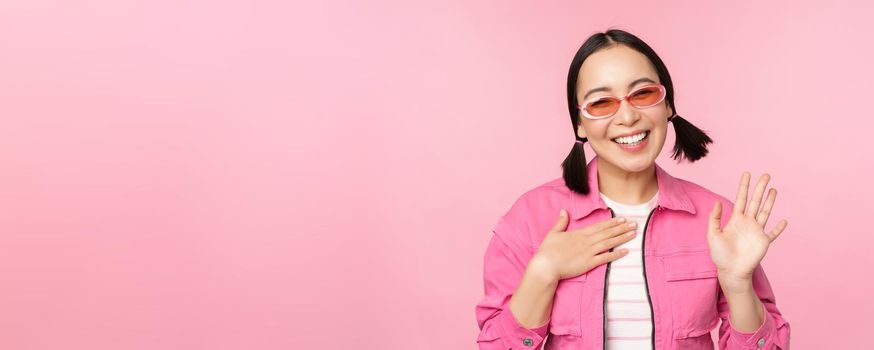 Portrait of friendly asian teen girl in sunglasses saying hello, waves her hand and smiles, greets you, hi gesture, stands over pink background.