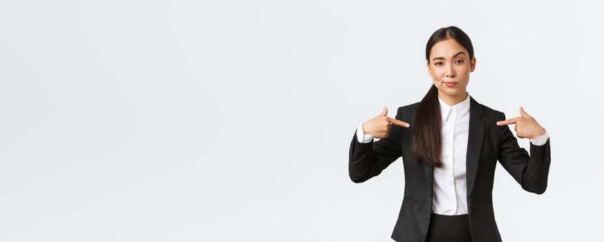 Professional asian businesswoman in suit pointing at herself as offering best deals. Proud and confident saleswoman show-off, suggest personal help or assistance, standing white background.