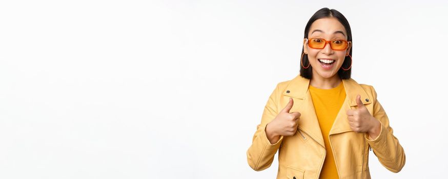 Enthusiastic asian girl smiles, shows thumbs up and looks happy, saying yes, approve and recommend smth, standing over white background.