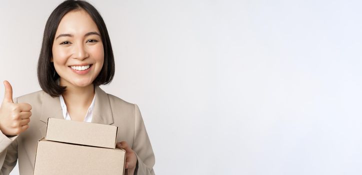 Smiling asian businesswoman, showing thumbs up and boxes with delivery goods, prepare order for client, standing over white background.