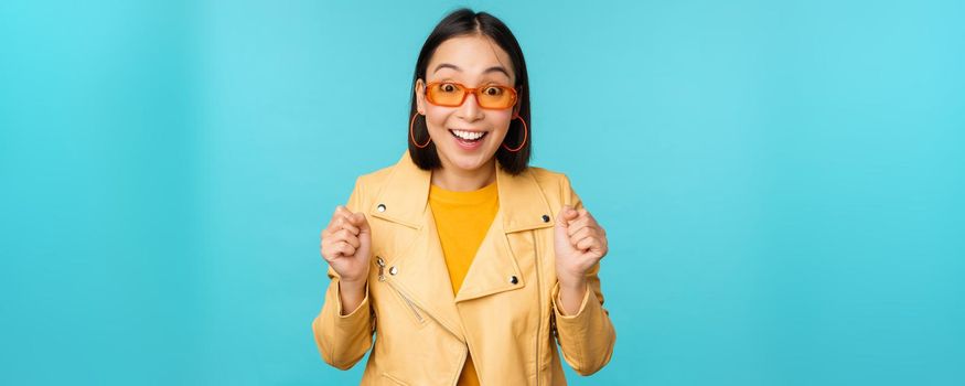 Image of enthusiastic young asian woman celebrating, triumphing, looking surprised and happy, clapping hands satisfied, standing over blue background.