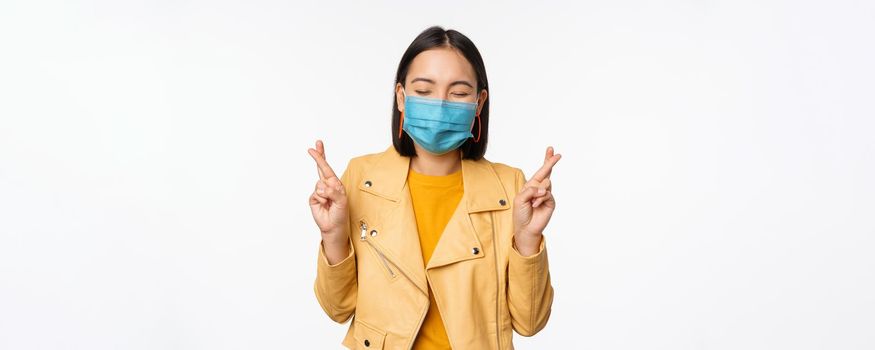 Hopeful asian girl in medical face mask, cross fingers, making wish, hoping, praying for smth, standing with anticipation over white studio background.