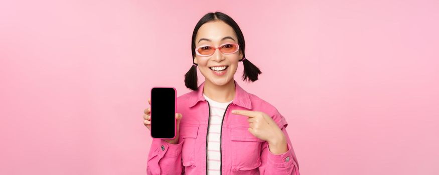 Enthusiastic asian woman in stylish clothes, sunglasses, pointing finger at mobile phone screen, showing smartphone application, standing over pink background.