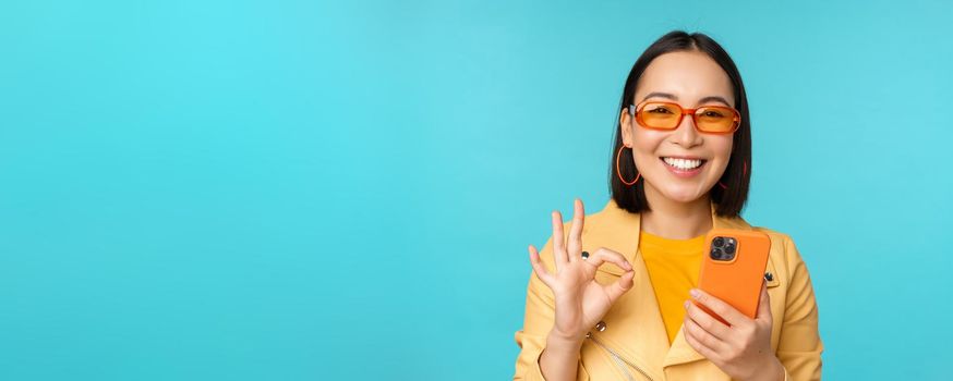 Happy korean girl in sunglasses, showing okay sign and holding mobile phone, using smartphone app, recommending application, standing over blue background.