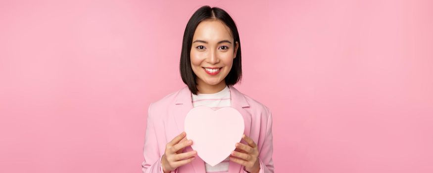 This is for you. Romantic cute asian corporate woman, girl in suit, showing heart-shaped box with gift, standing over pink background.